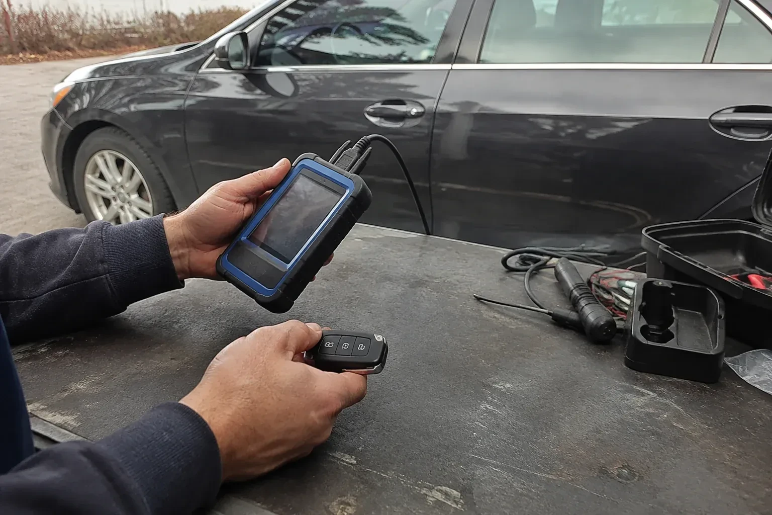 Hands holding a blue-edged electronic device and car-key fob at a worktable beside a parked car. — Transponder Key Programming, Cambridge